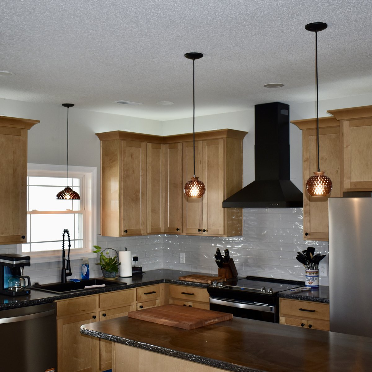 Three handmade bloodwood pendant lights in a row over a kitchen with dark granite countertops and maple cabinets